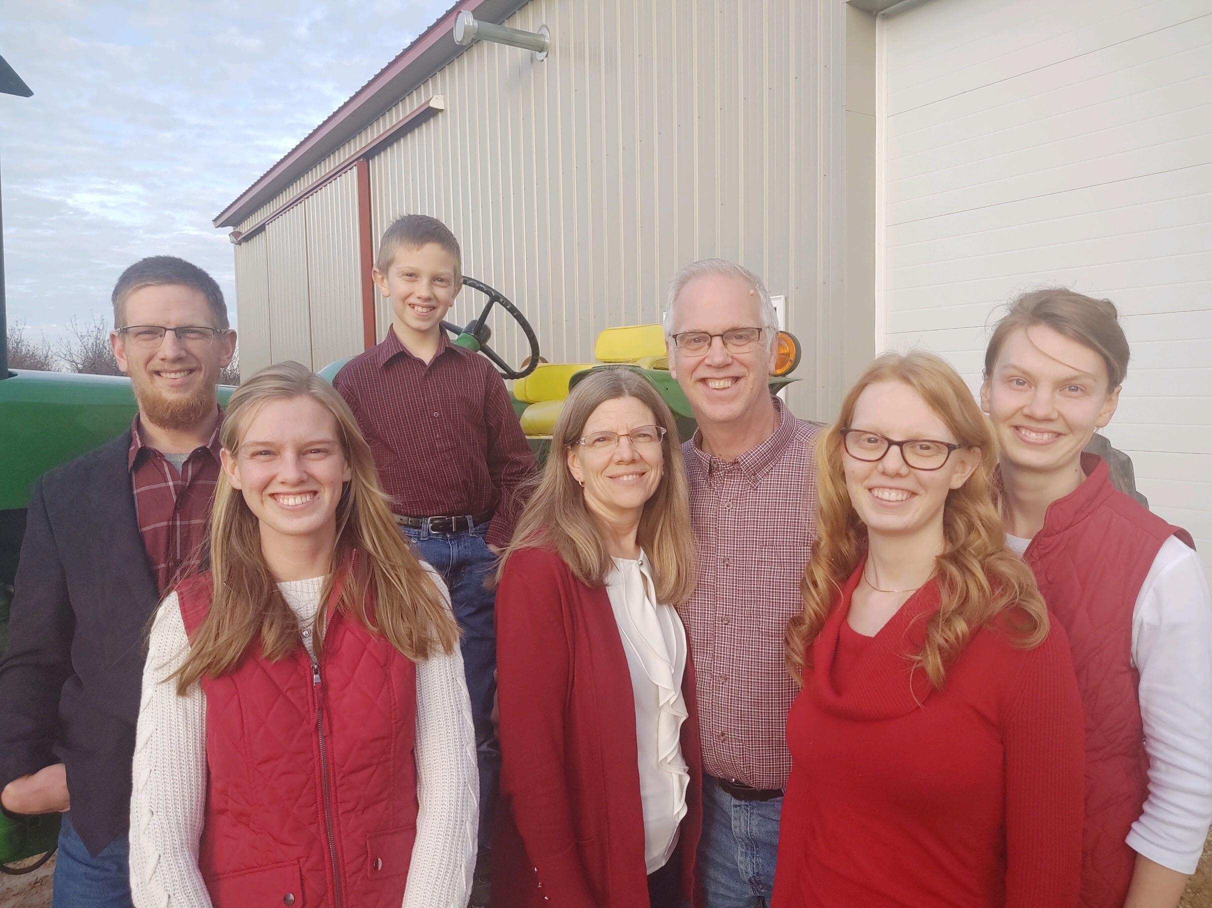 A happy family of seven poses outdoors in front of a tractor, wearing coordinated outfits in shades of red and plaid. The setting features a barn-like structure, highlighting a rural lifestyle.