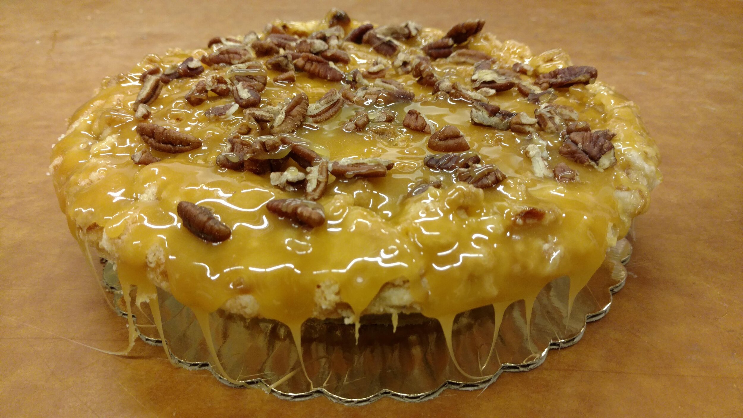 Pecan pie topped with a glossy caramel glaze and sprinkled with chopped pecans, presented on a silver pie dish against a wooden background.