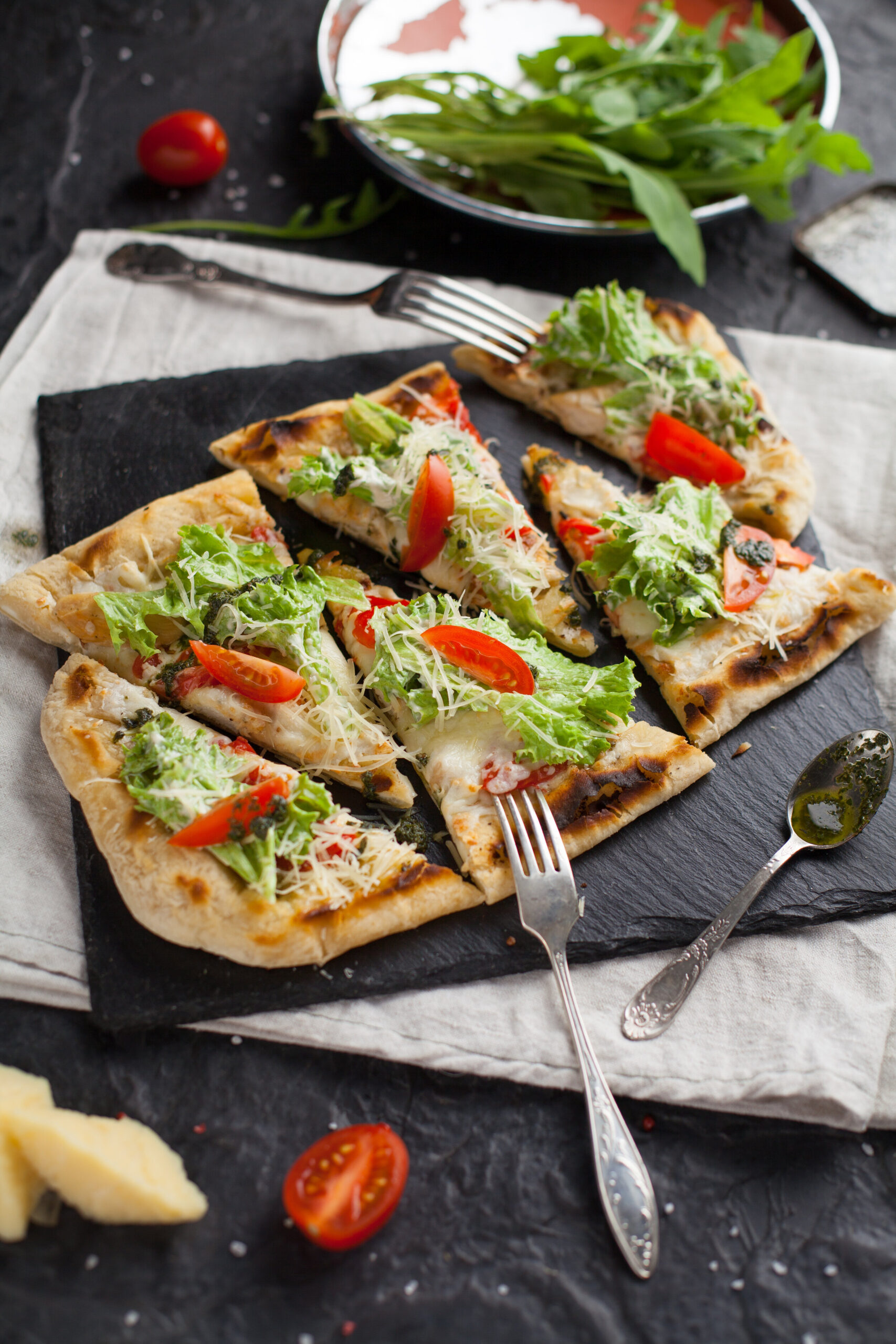Delicious flatbread pizza topped with fresh lettuce, sliced tomatoes, and shredded cheese, served on a black slate board with forks and a side of salad.